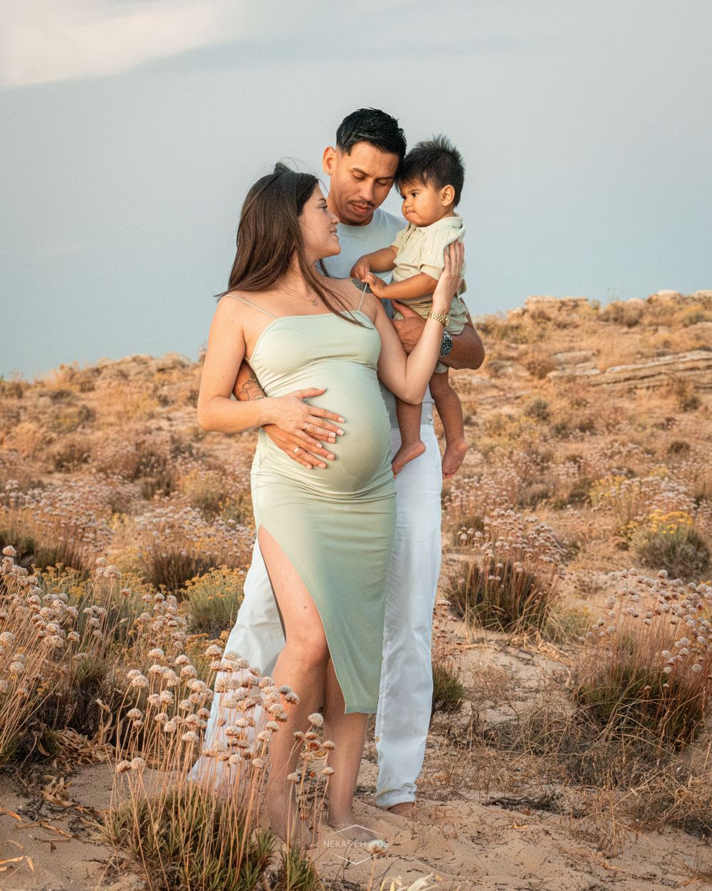 Pregnant woman in mint dress with partner holding toddler, posing outdoors in desert landscape for maternity photo.