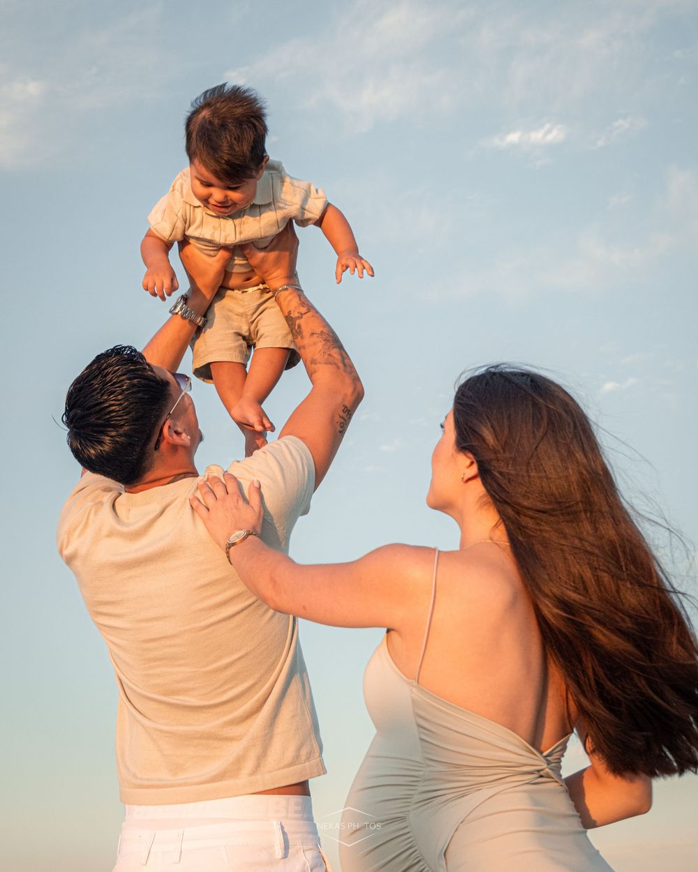 Parents joyfully lifting their baby in the air against a blue sky at golden hour on a sunny day.