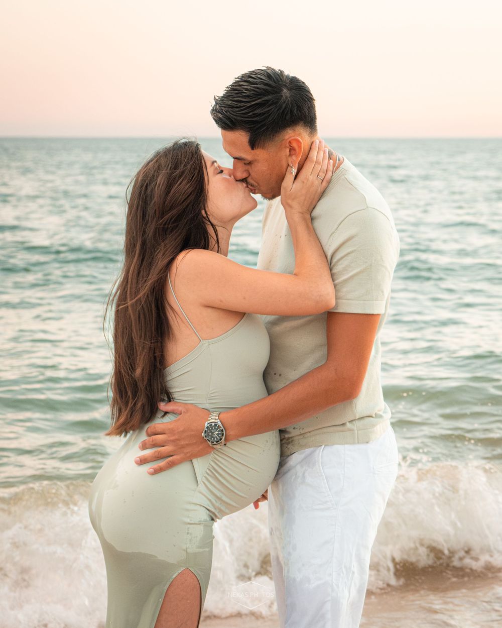 Couple sharing a romantic kiss on the beach at sunset, woman in sage green dress, man in matching outfit.