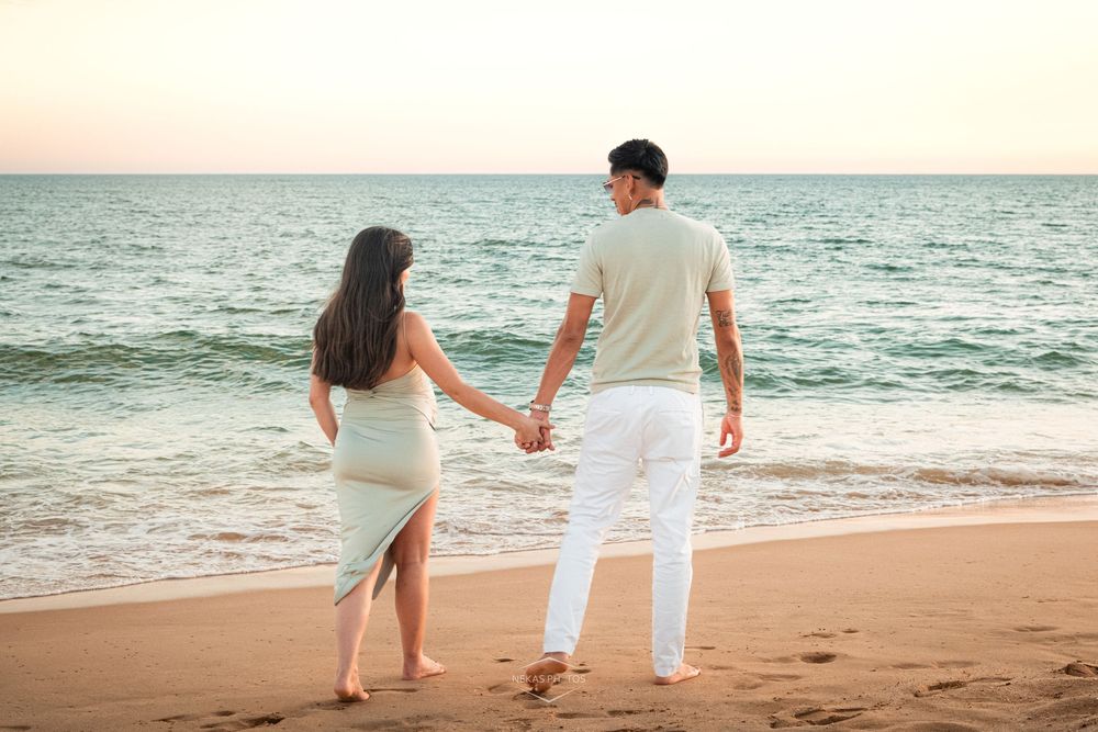 Couple holding hands on a sandy beach at sunset, facing the ocean in coordinated light-colored outfits.