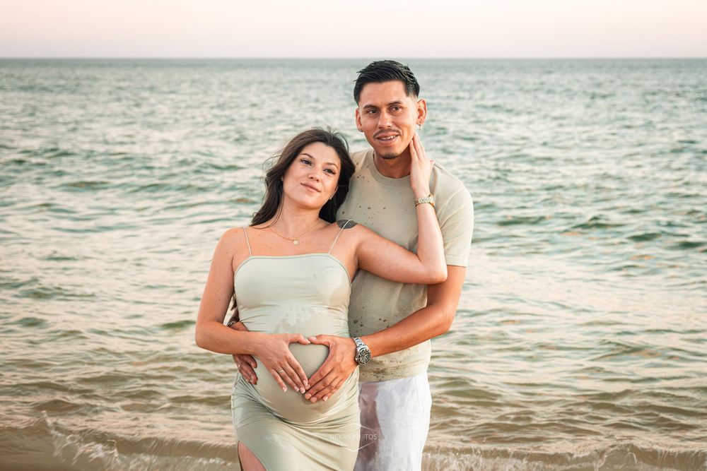 Pregnant couple posing on beach at sunset, woman in sage green dress with partner forming heart shape on baby bump.
