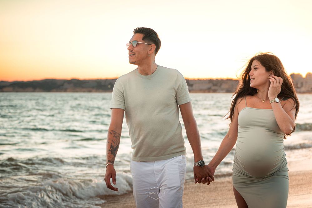Pregnant couple holding hands walking on beach at sunset, man in sage shirt and white pants, woman in gray maternity dress.