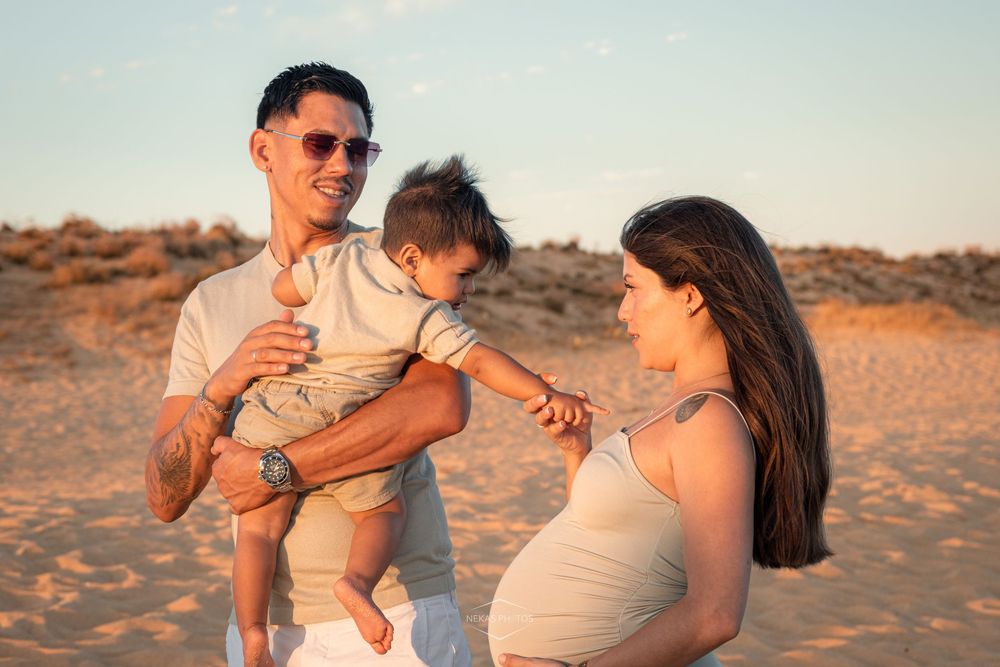 Pregnant woman and man holding toddler on sandy beach at golden hour sunset.
