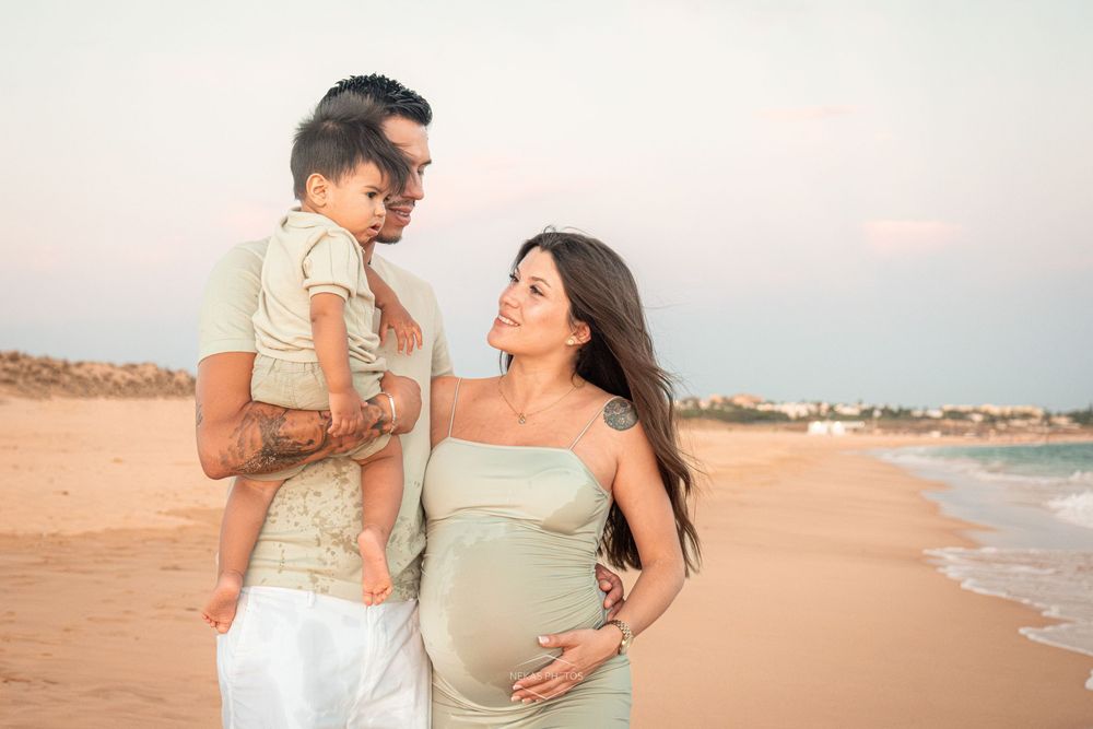 Pregnant woman in green dress with partner and child on beach at sunset.