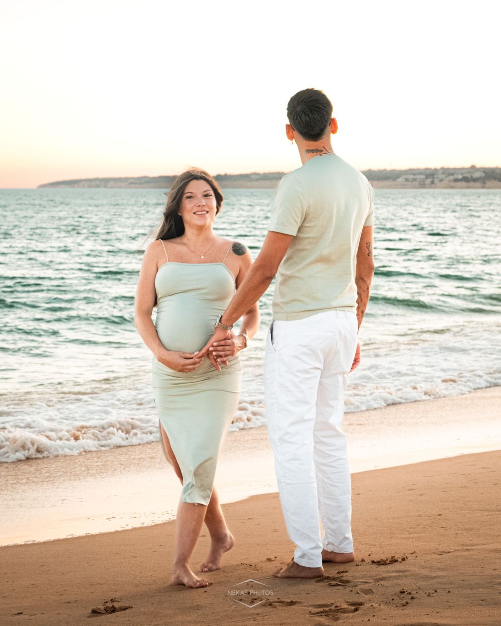 Pregnant couple holding hands on a sandy beach at sunset, woman in sage green dress cradling baby bump.
