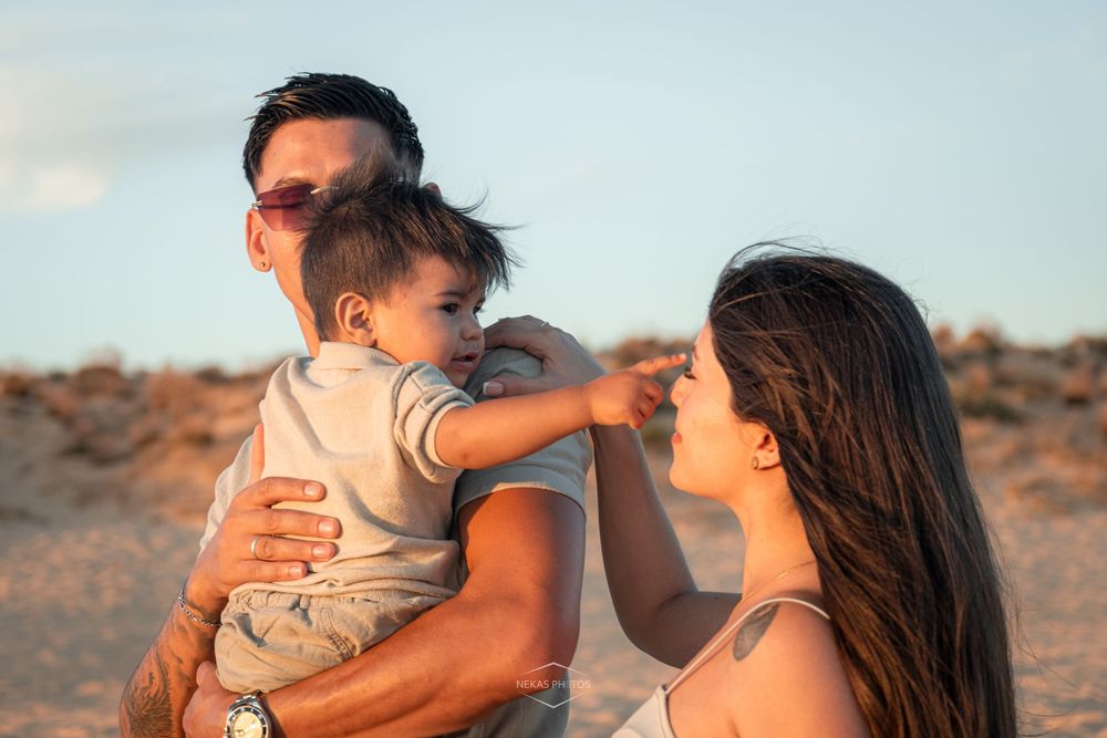 A young family with a baby enjoys a sunny beach day, with the child reaching toward the mother.