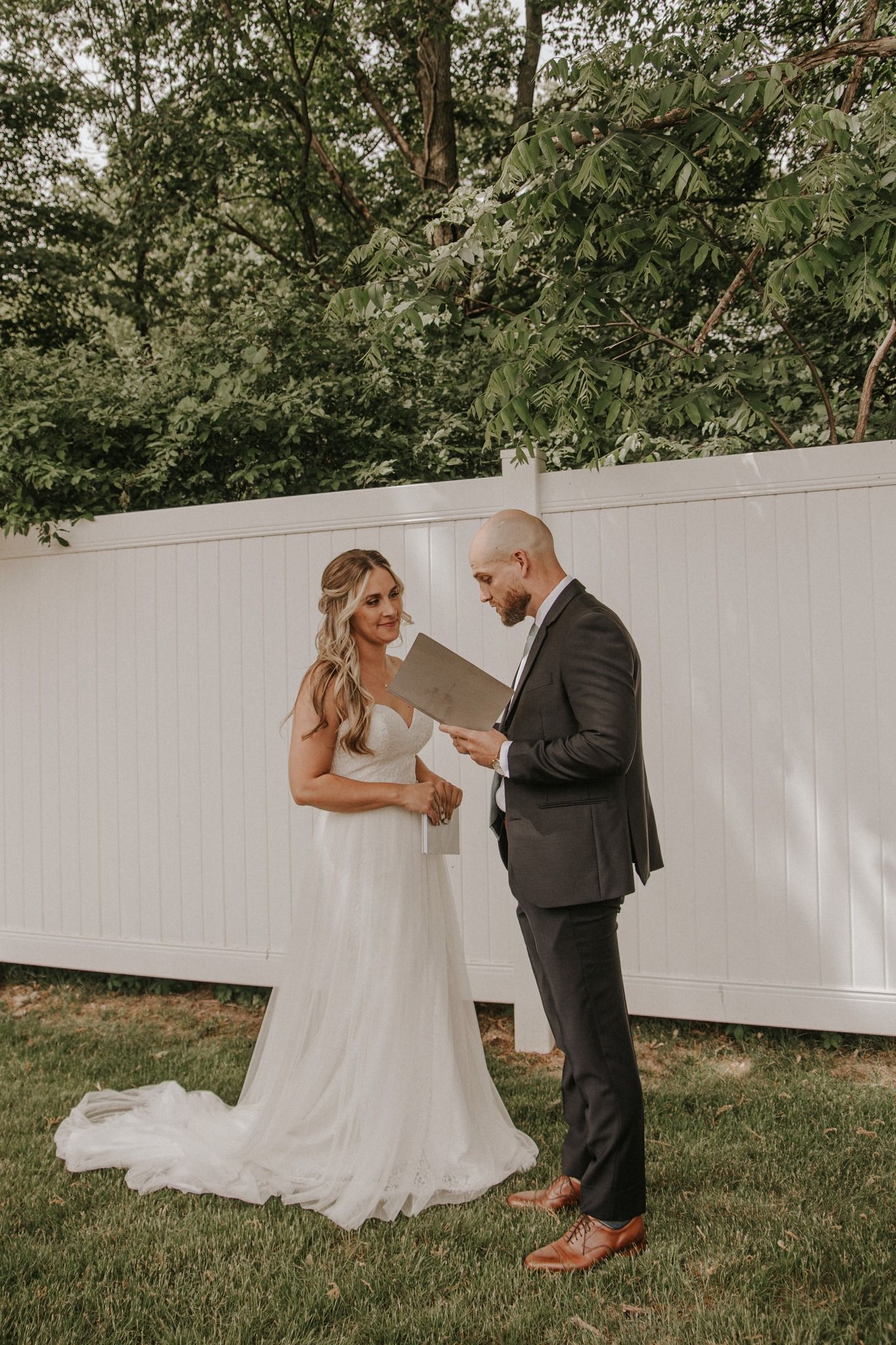 Bride in white gown and groom in dark suit exchange vows outdoors, reading from a booklet in a garden setting.