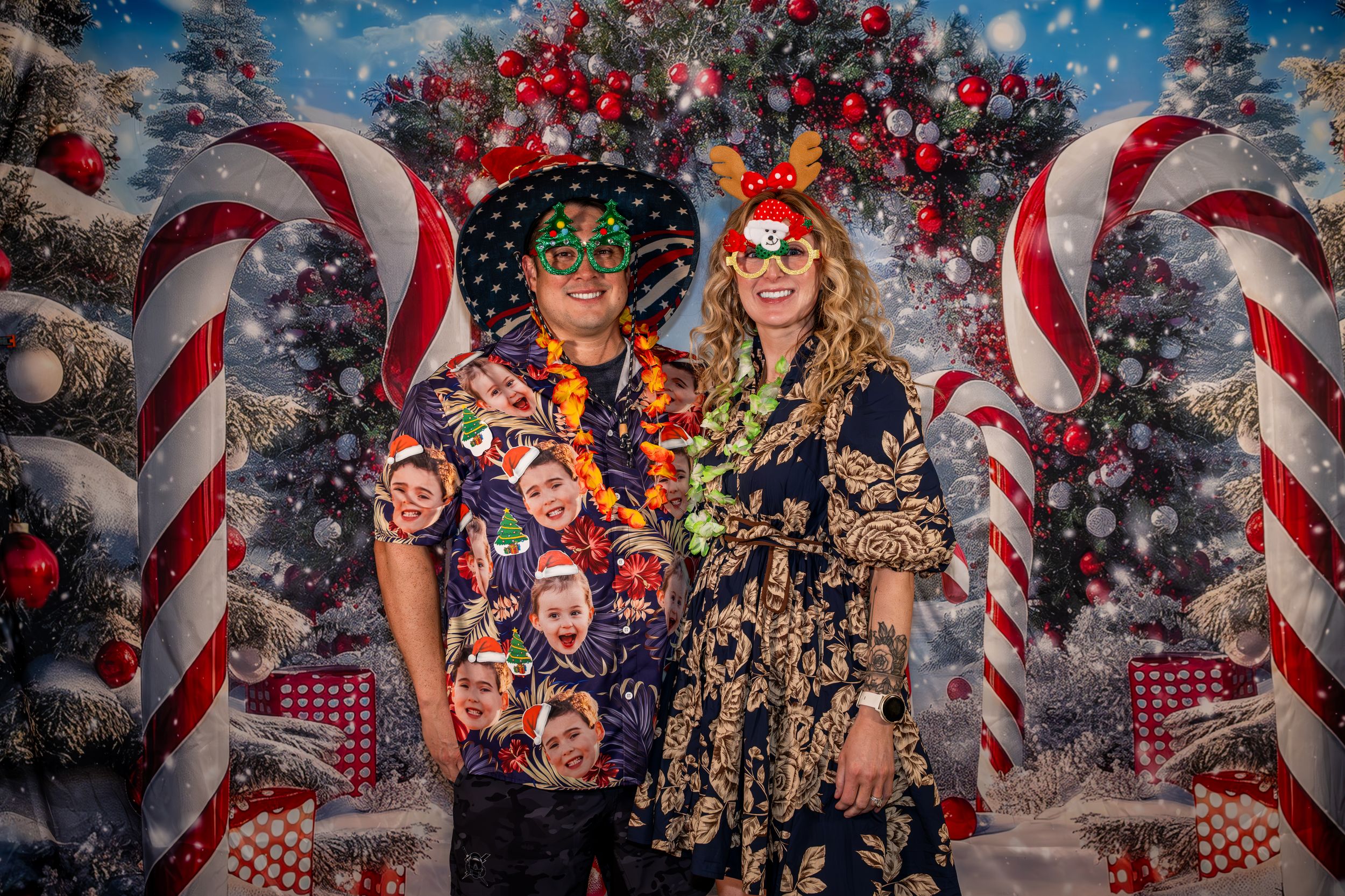 Couple wearing festive Hawaiian shirts and holiday accessories posing in front of a Christmas-themed backdrop with candy canes.