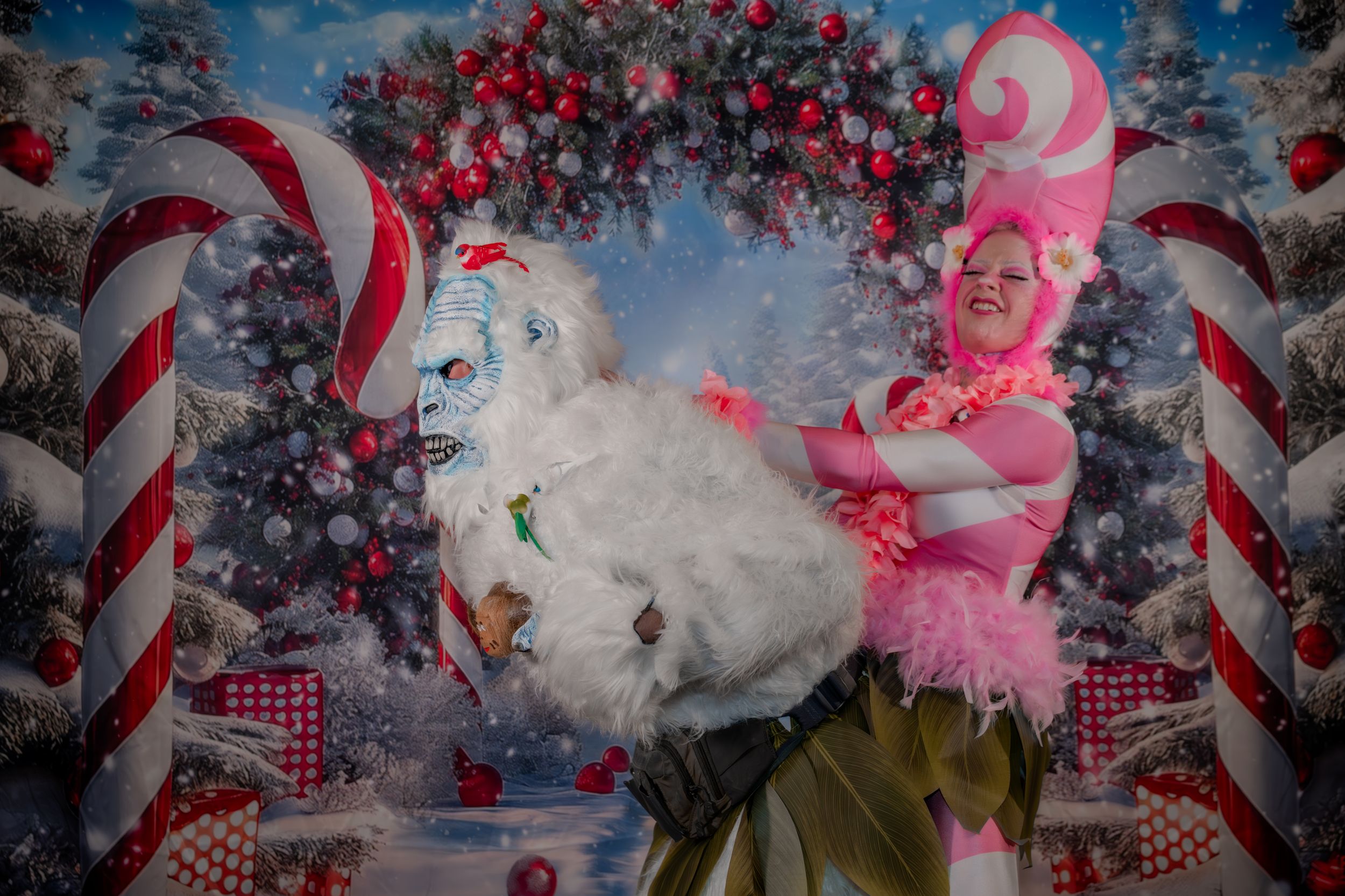 Two performers in colorful Christmas costumes pose in front of a festive holiday backdrop with candy canes and ornaments.