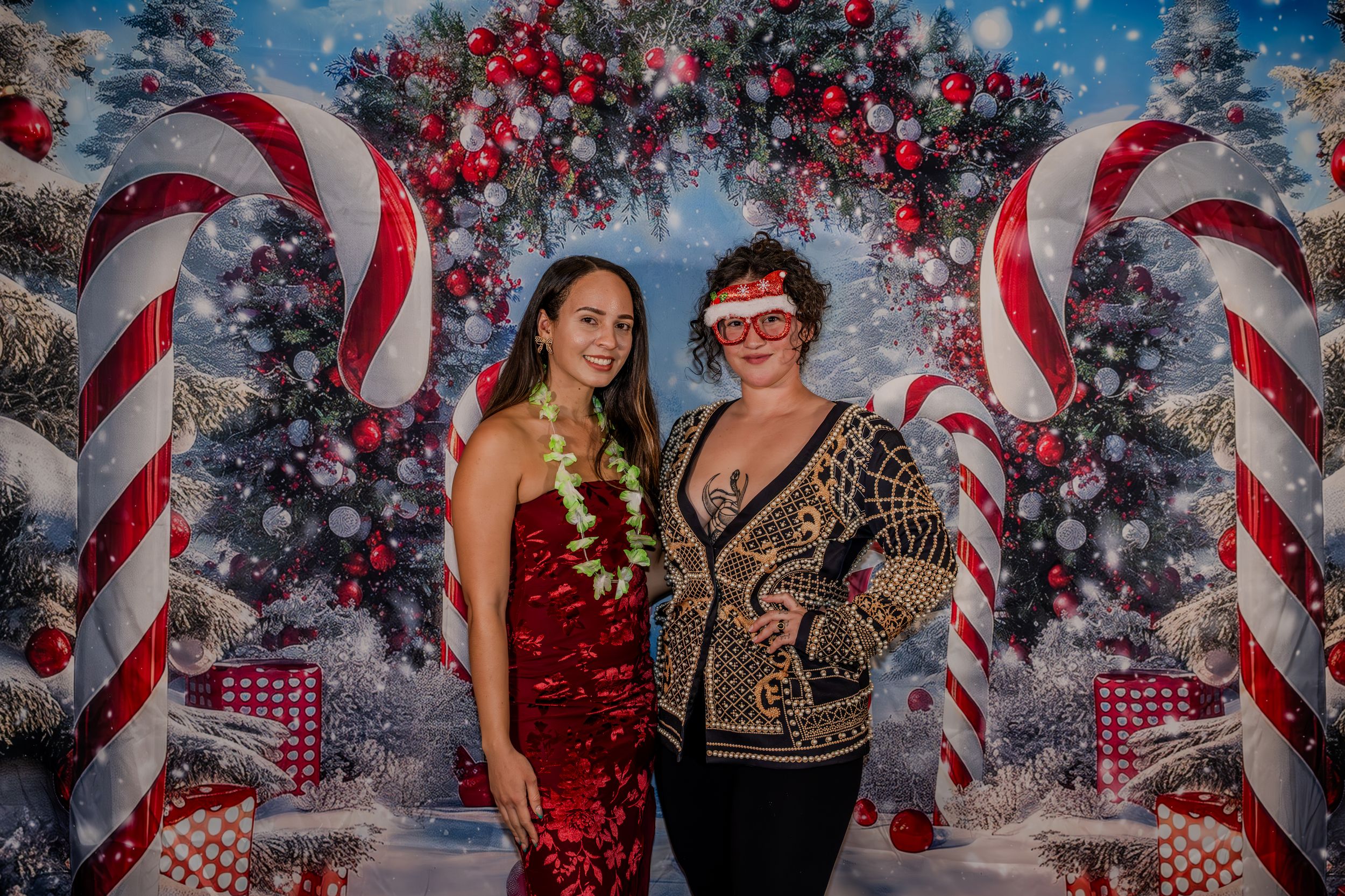 Two women posing in front of a festive Christmas backdrop with candy canes and ornaments at a holiday party.