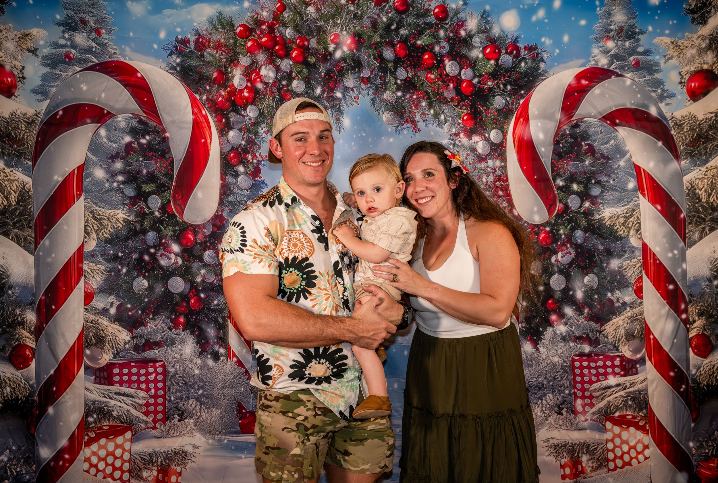 Family of three posing in front of festive Christmas backdrop with candy canes and decorated tree.