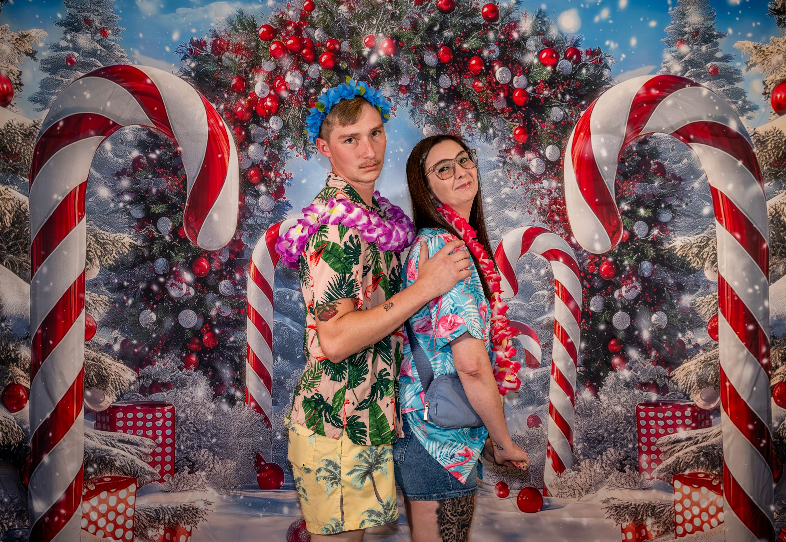 Two people in tropical Hawaiian shirts pose back-to-back in front of a festive Christmas backdrop with candy canes.