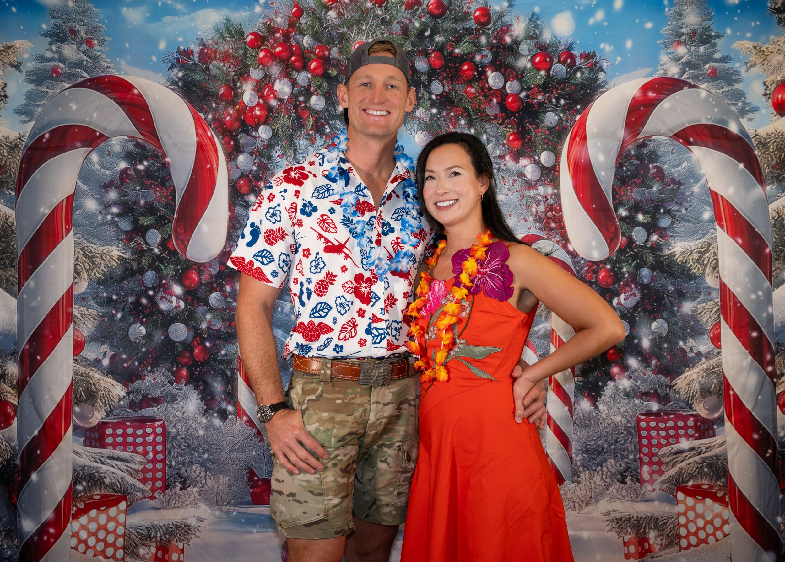 Couple posing in front of festive Christmas backdrop with candy canes and ornaments, wearing tropical holiday attire.