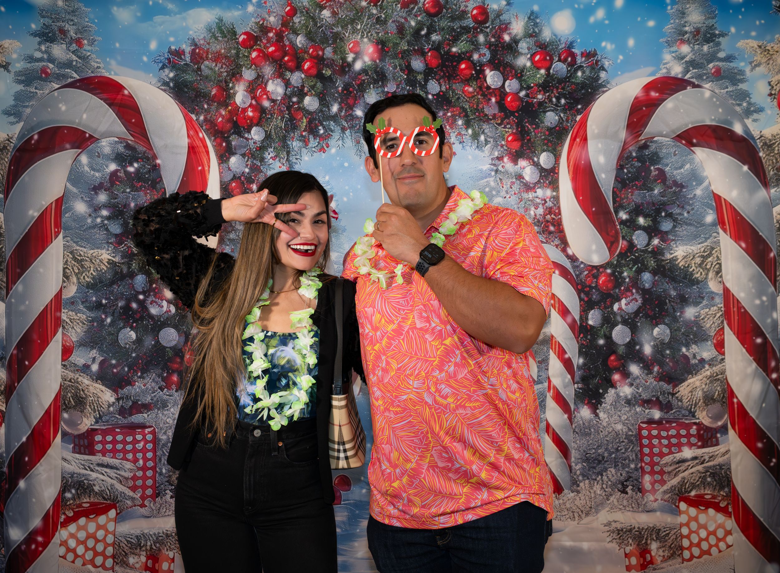 Two people pose in tropical attire before a festive Christmas backdrop with candy canes and decorated trees.