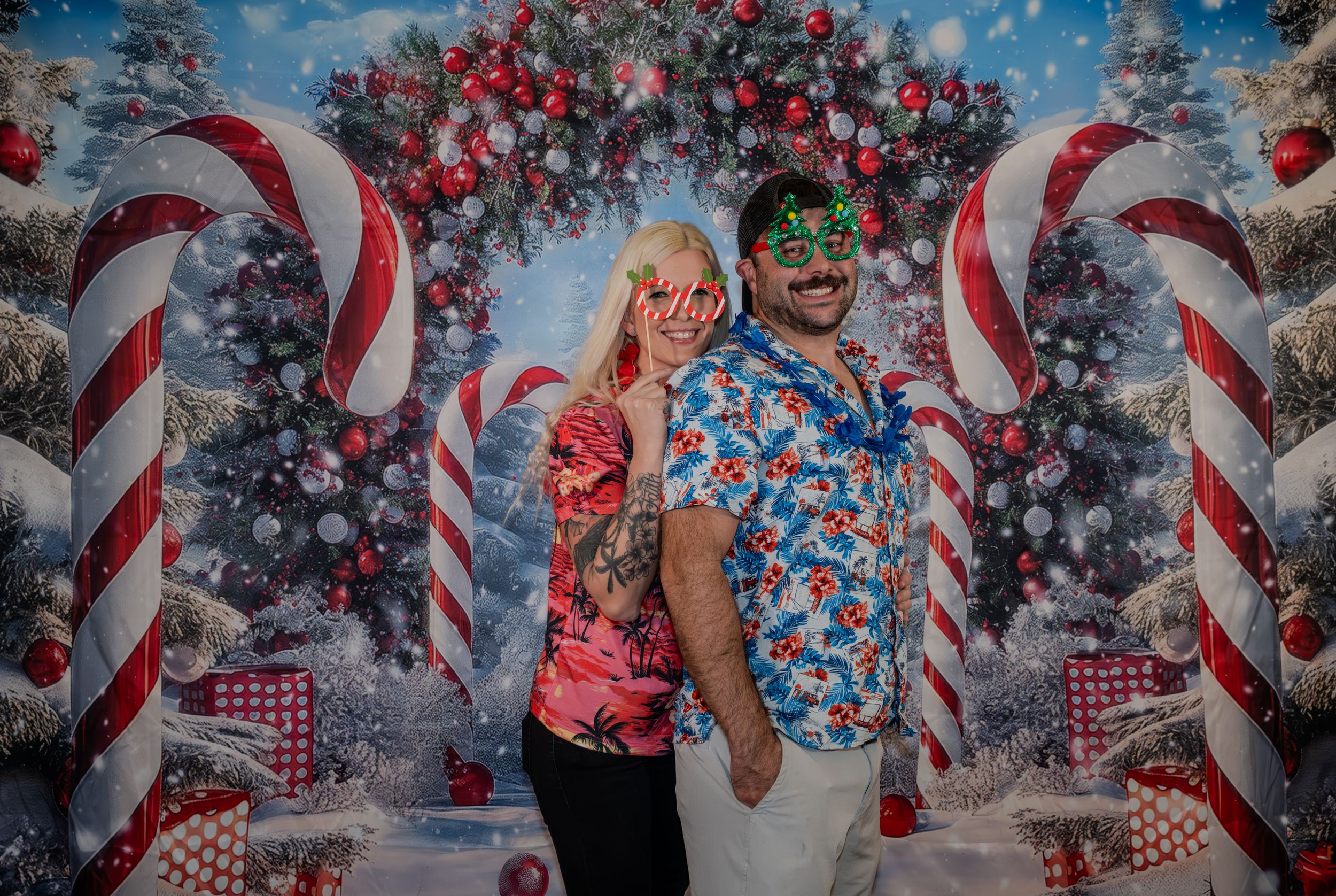 Couple posing in tropical Christmas shirts in front of festive candy cane and Christmas tree holiday backdrop.