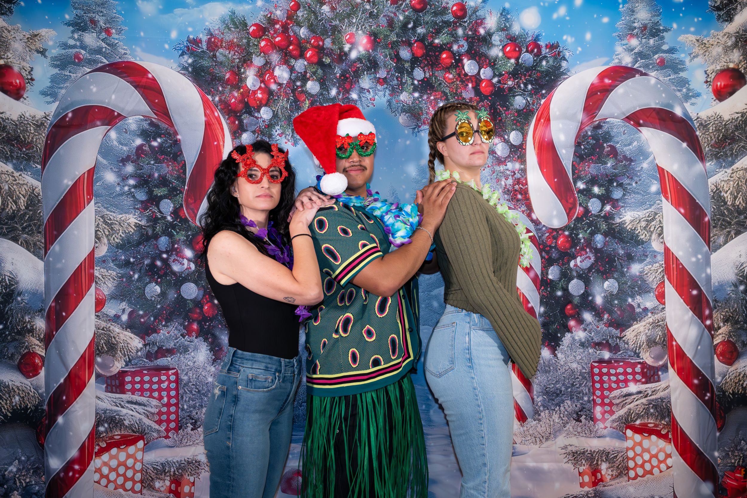 Three women pose in festive Christmas photo booth with candy canes, holiday trees, and wearing fun seasonal accessories.