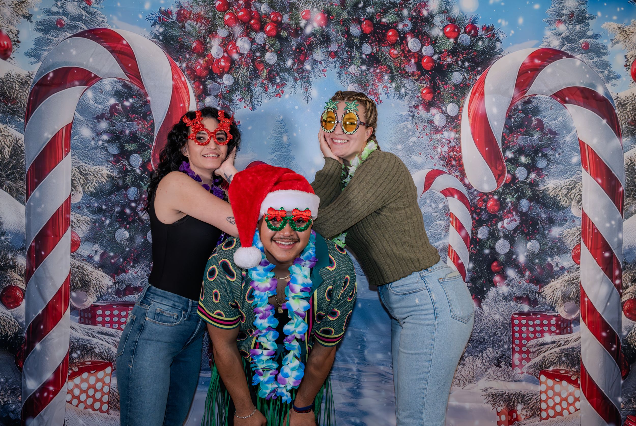 Three friends wearing fun Christmas accessories posing before a festive candy cane backdrop.