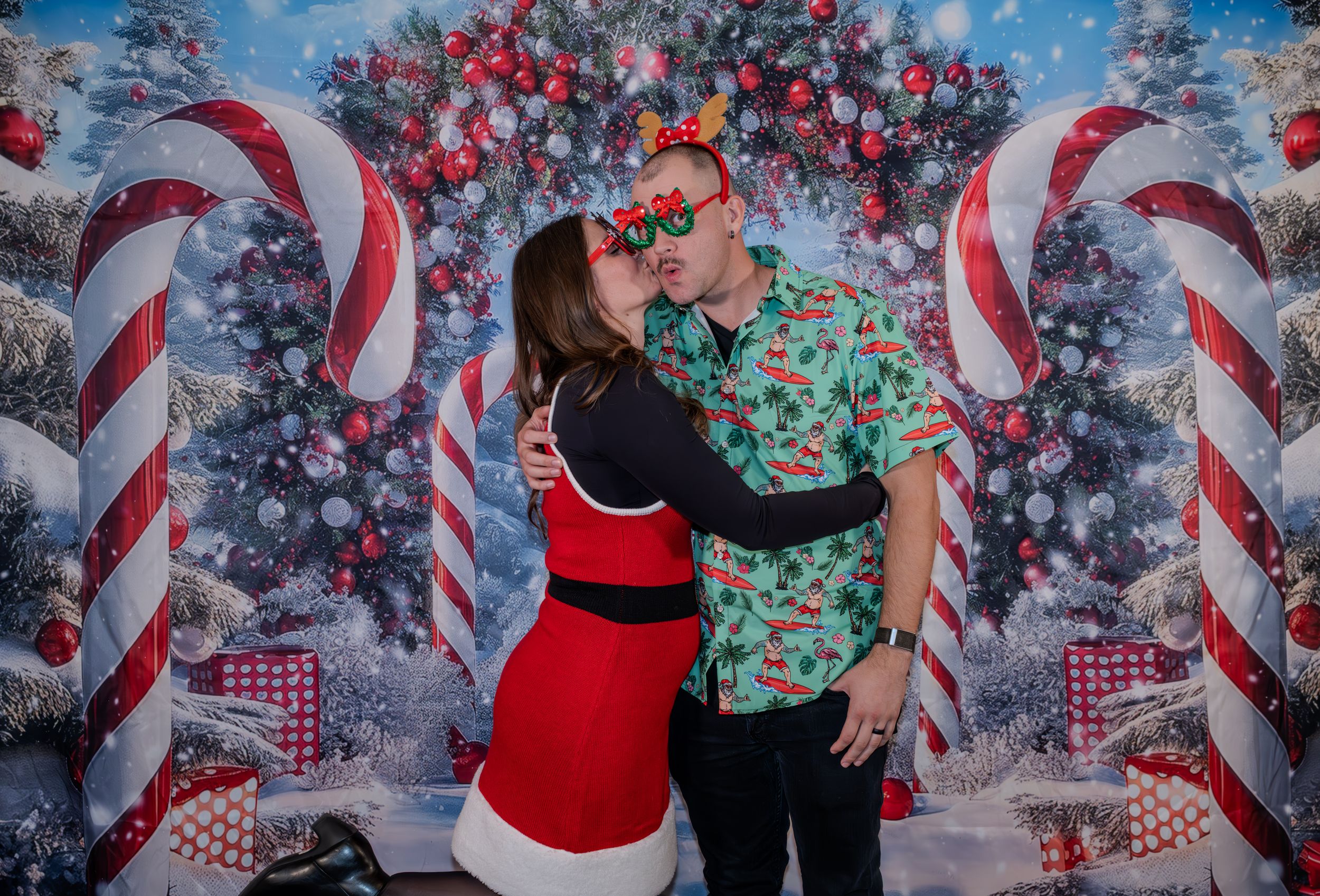 Couple kissing in front of festive Christmas backdrop with candy canes, wearing holiday outfits and reindeer antler glasses.