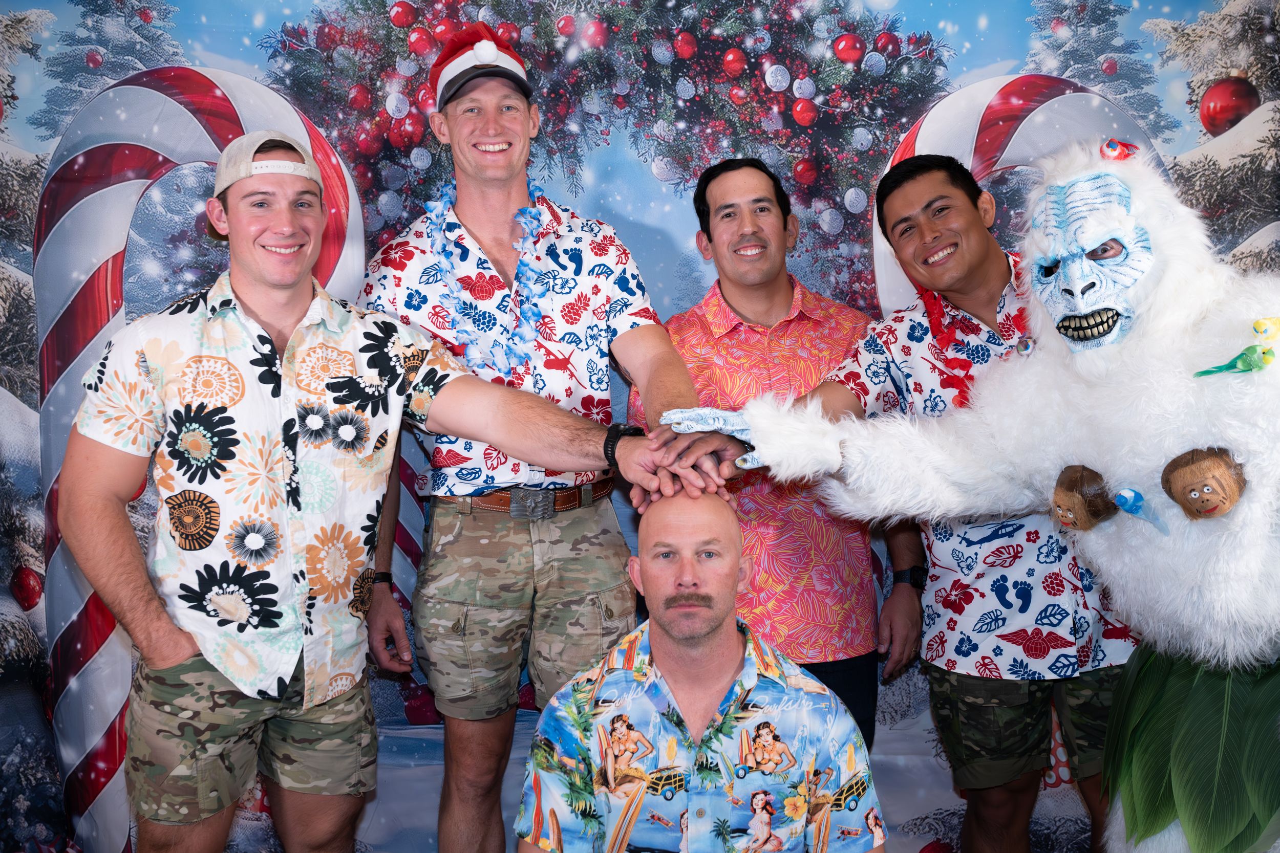 Five men in tropical Christmas shirts pose with a yeti mascot in front of a festive holiday backdrop.