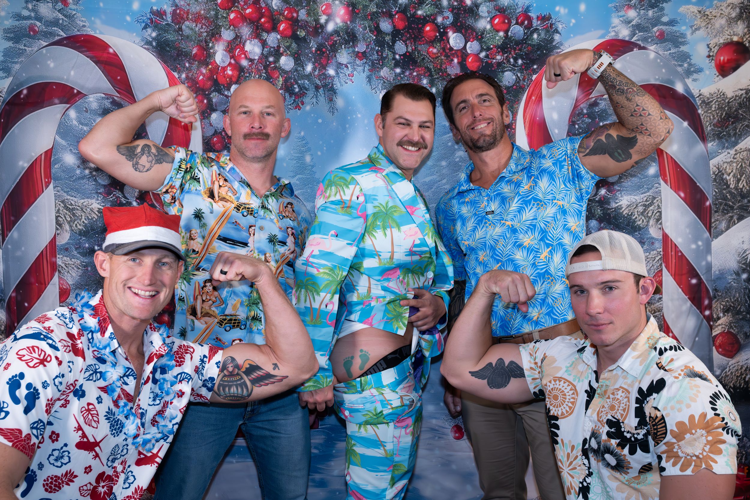 Five men in tropical Hawaiian shirts pose playfully in front of a festive Christmas holiday backdrop with candy canes.