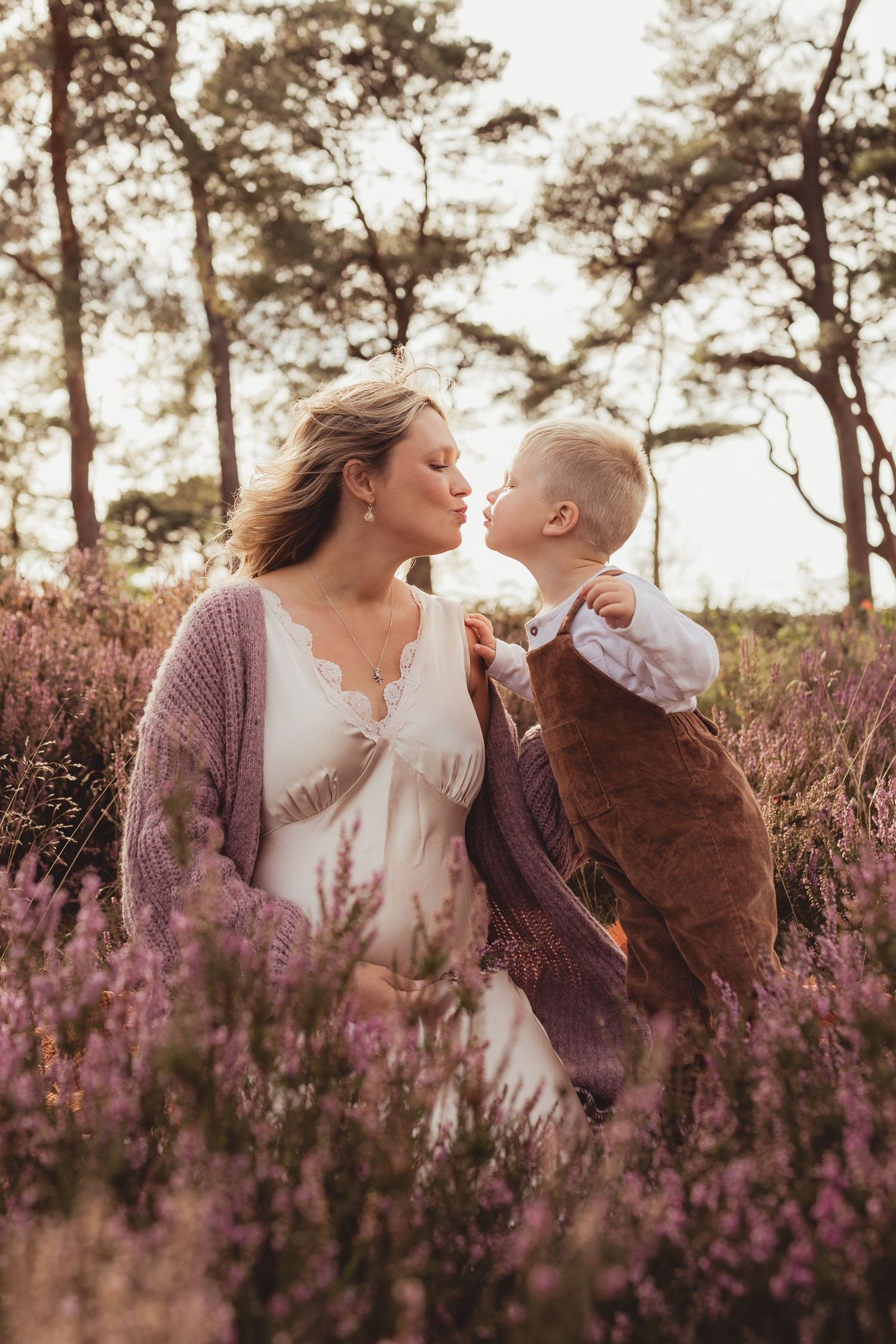 A mother and toddler share a sweet kiss in a purple heather field, surrounded by trees in warm golden light.