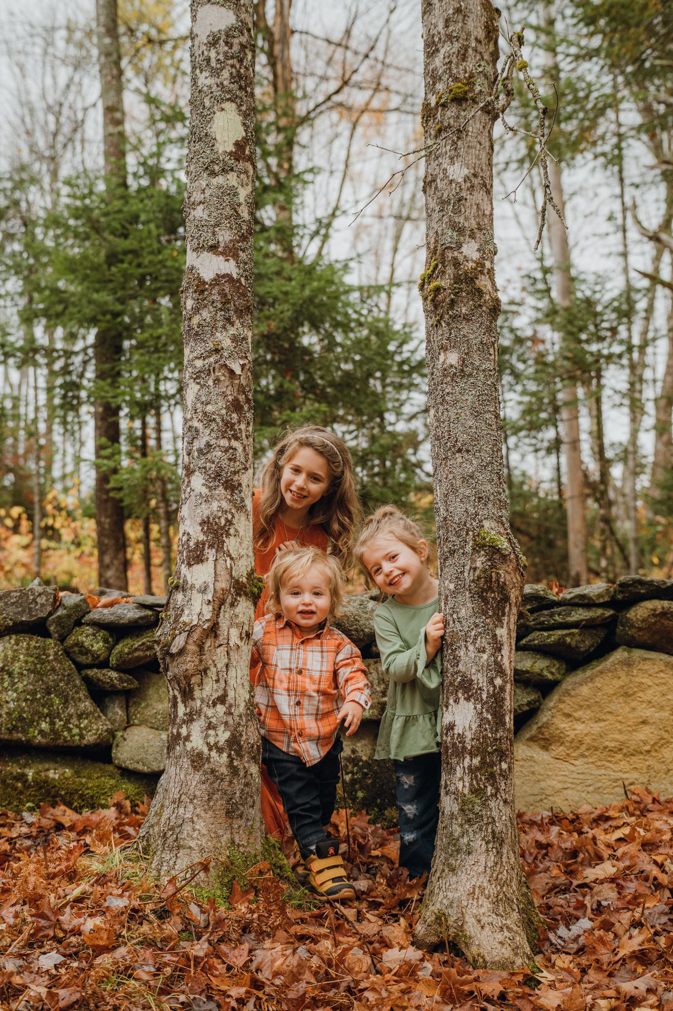 Three children pose between two trees in an autumn forest with fallen leaves and a stone wall backdrop.
