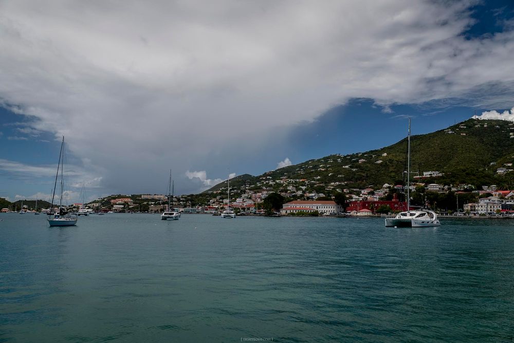 Sailboats anchored in calm turquoise harbor with colorful hillside town and dramatic cloudy sky backdrop.