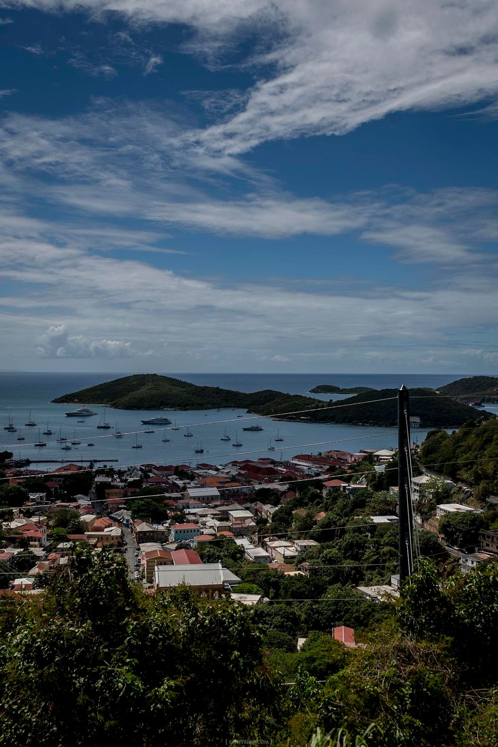 Aerial view of a Caribbean coastal town with colorful rooftops, sailboats in a turquoise bay, and lush green hills under blue skies.