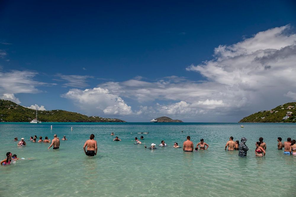 Tourists swimming in crystal-clear turquoise Caribbean waters with lush green hills and dramatic clouds in the background.