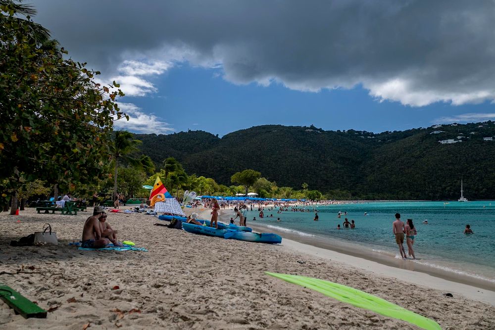 Tropical beach scene with kayaks, sunbathers, and calm turquoise waters surrounded by lush green hills under cloudy skies.