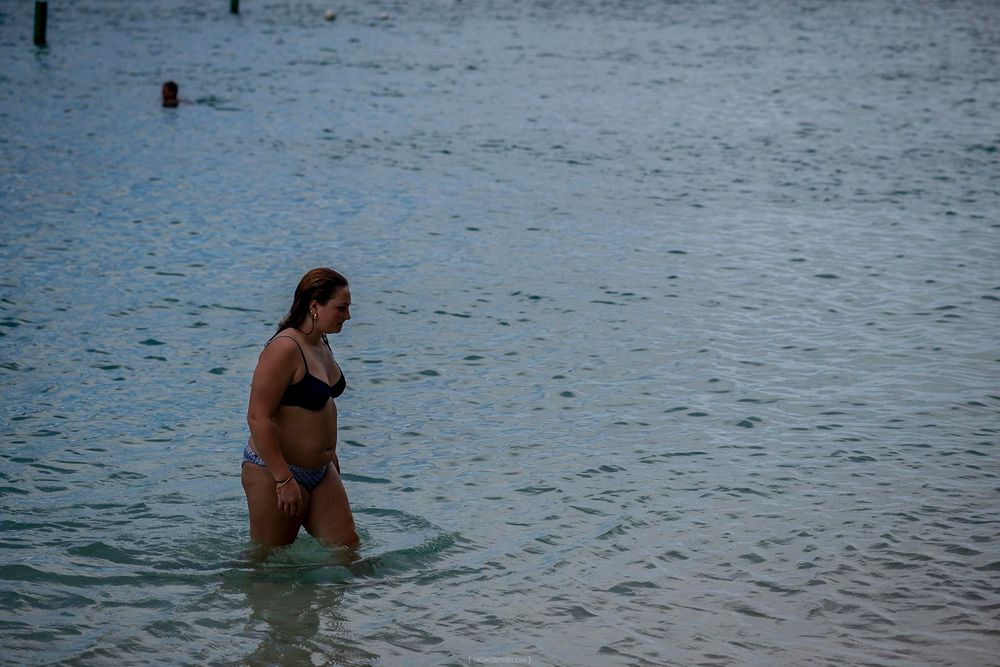 Woman in black bikini wading in calm shallow ocean water near a beach.