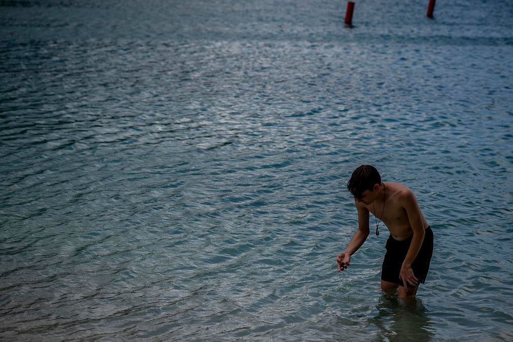 Shirtless man wading in calm shallow ocean water, bending forward to touch the sea surface.