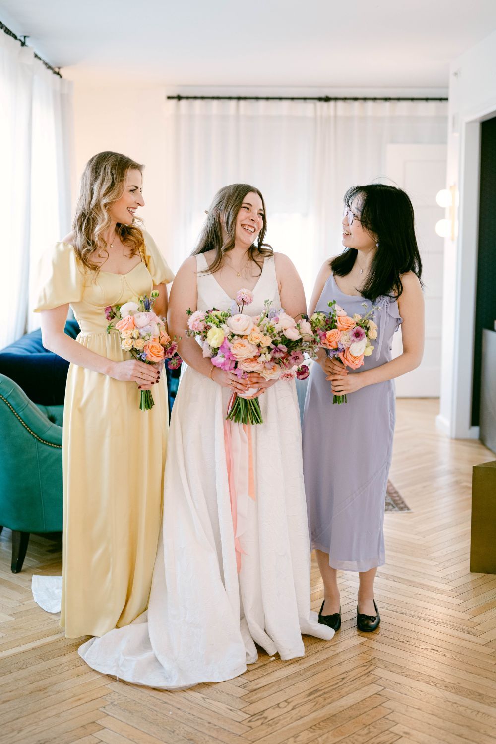 Bride in white gown stands with two bridesmaids in yellow and lavender dresses holding colorful bouquets indoors.