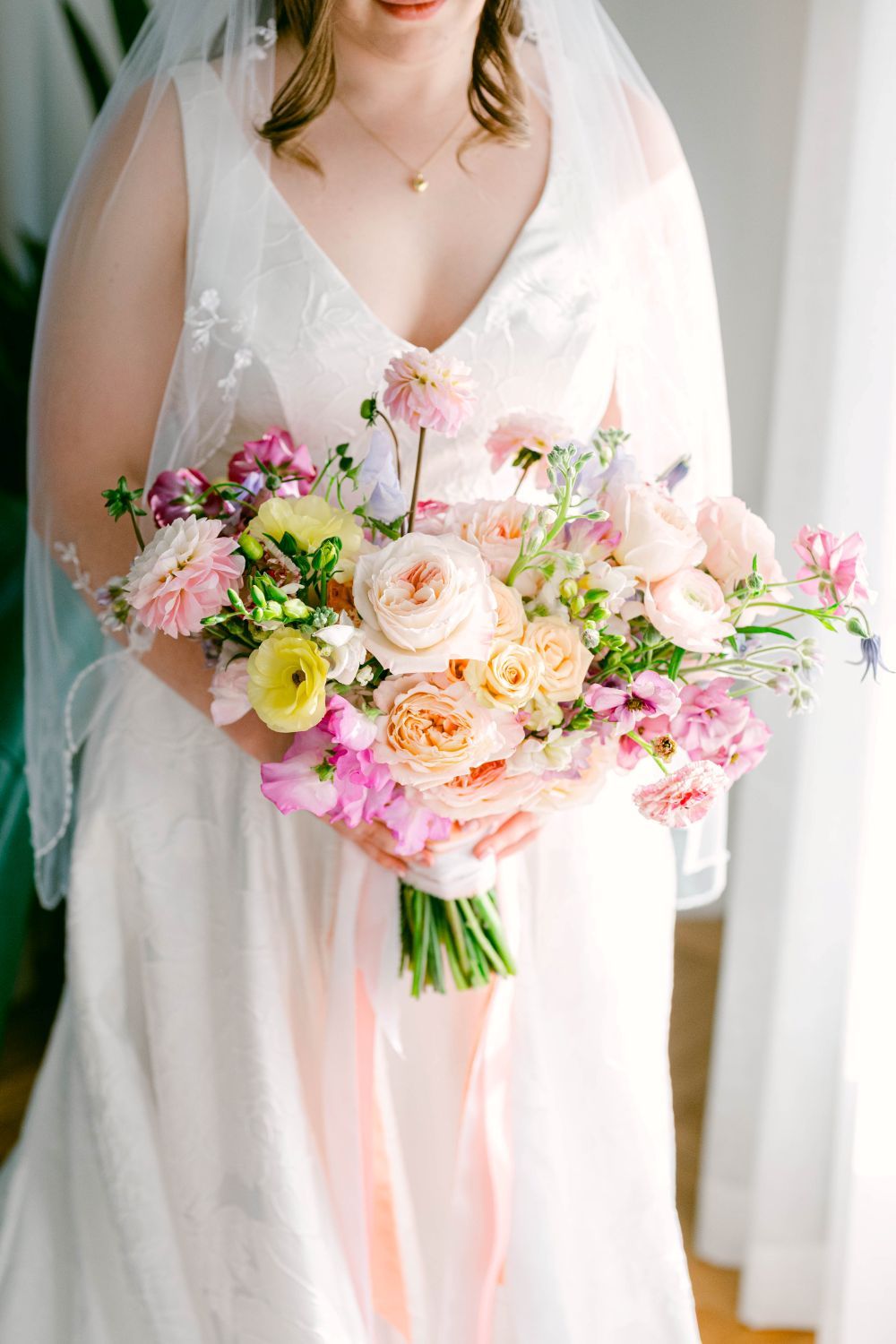 Bride in white lace gown holding a lush pink, peach, and yellow floral wedding bouquet with flowing ribbons.