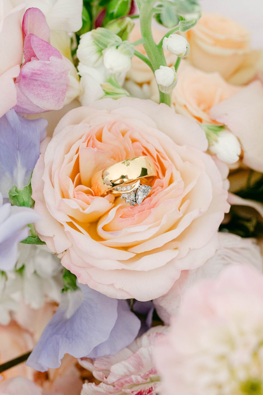 Gold wedding bands and diamond engagement ring nestled in a blush peach garden rose bouquet with lavender and white flowers.