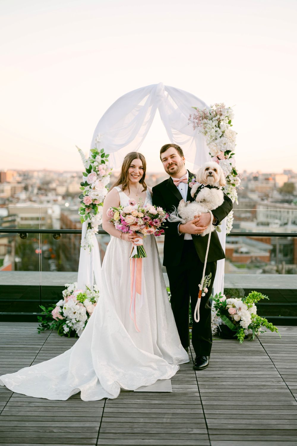 Bride and groom pose with small white dog under floral arch on rooftop at sunset.