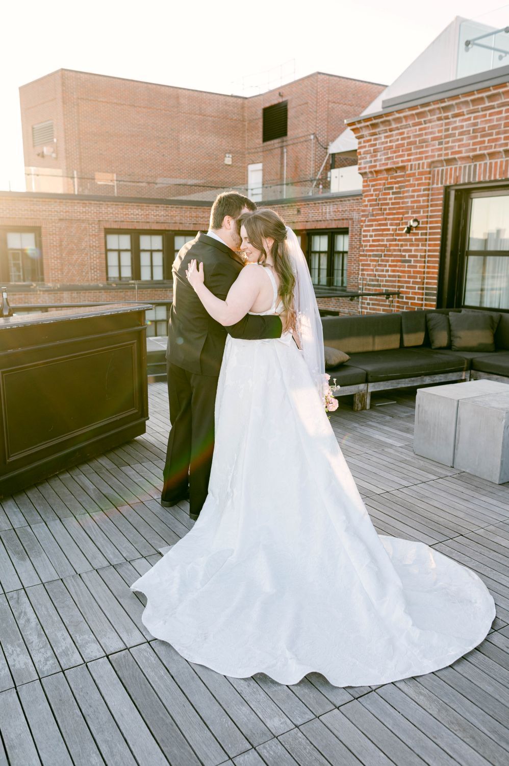 Bride and groom share a romantic embrace on a rooftop deck at golden hour, with brick buildings in the background.