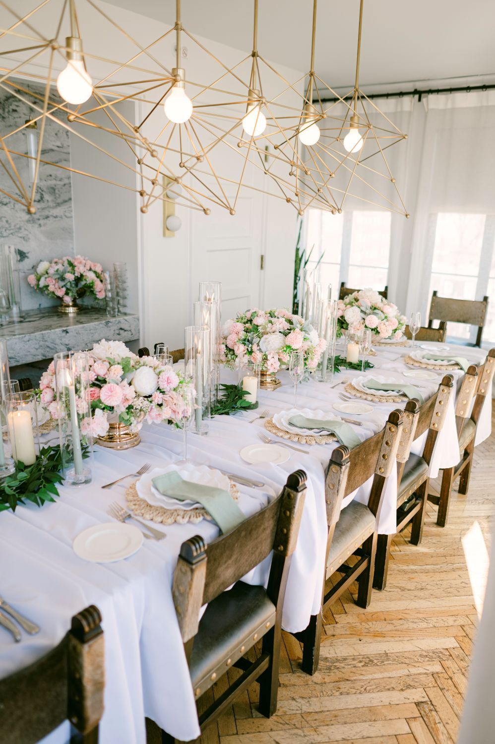 Elegant wedding reception table with pink floral centerpieces, gold geometric chandeliers, and white linens on herringbone floors.