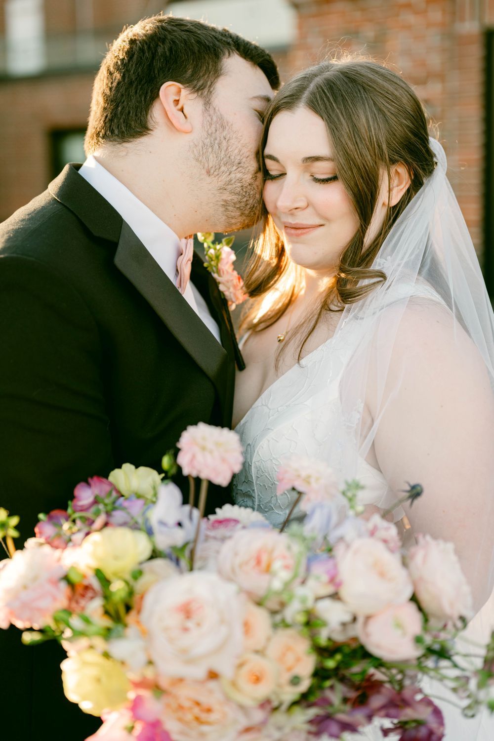 Groom kissing bride on cheek while she smiles holding colorful pink and yellow floral bouquet outdoors.