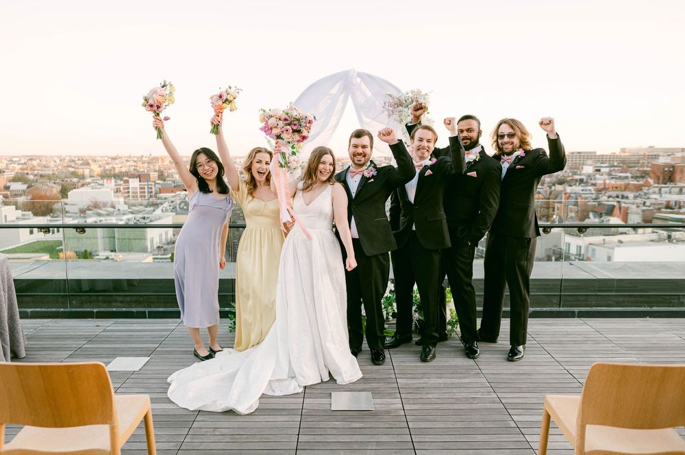 Wedding party celebrating on rooftop at sunset, bride in white gown, groom in tuxedo, bridesmaids and groomsmen cheering.