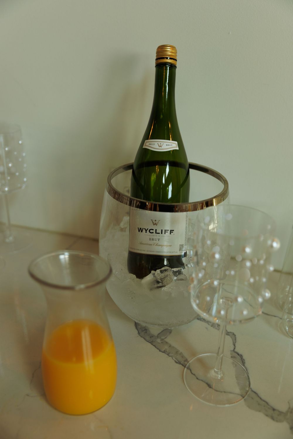 Wycliff champagne bottle chilling in ice bucket beside orange juice carafe and crystal glasses on marble surface.
