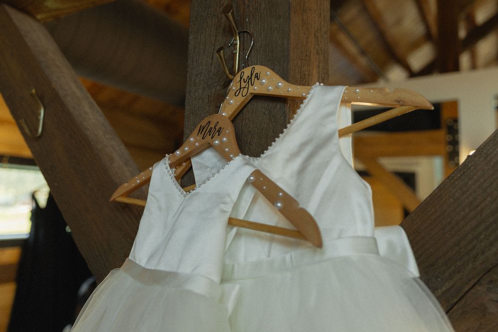 Two white flower girl dresses hanging on personalized wooden hangers in a rustic barn wedding venue.