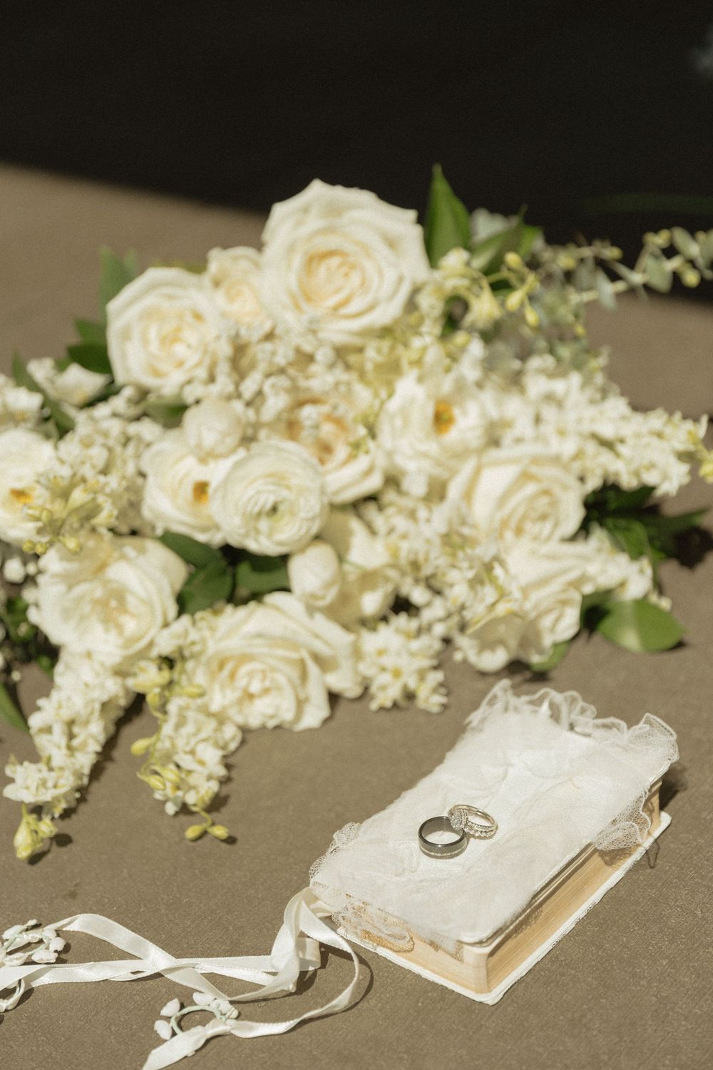 White roses bridal bouquet with wedding rings on lace ring bearer pillow, elegant wedding detail photography.