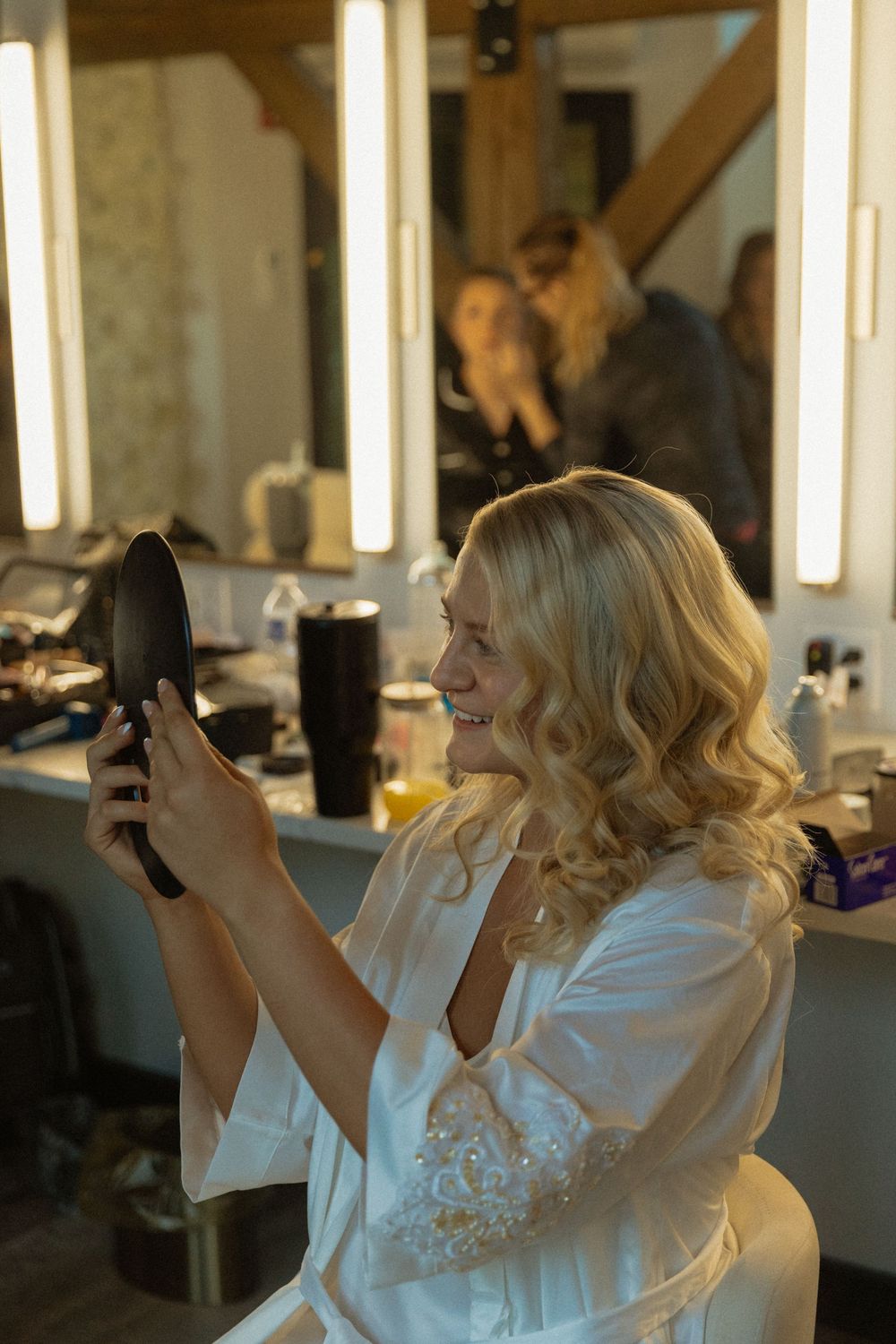 Smiling blonde bride in white robe admires her curled hair in a hand mirror at a bridal makeup vanity.