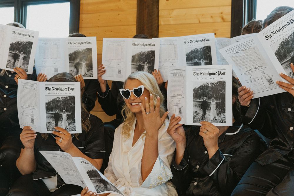 Bride in white robe and heart sunglasses poses with bridesmaids holding The Knot Post newspapers at wedding.