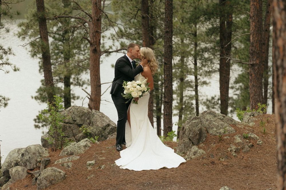 Bride and groom kissing among pine trees with a lake backdrop on their wedding day.