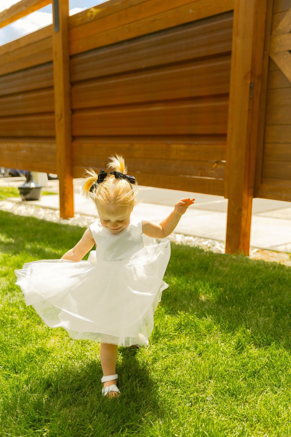 Adorable blonde toddler girl in white dress twirling on green grass at outdoor wedding venue.