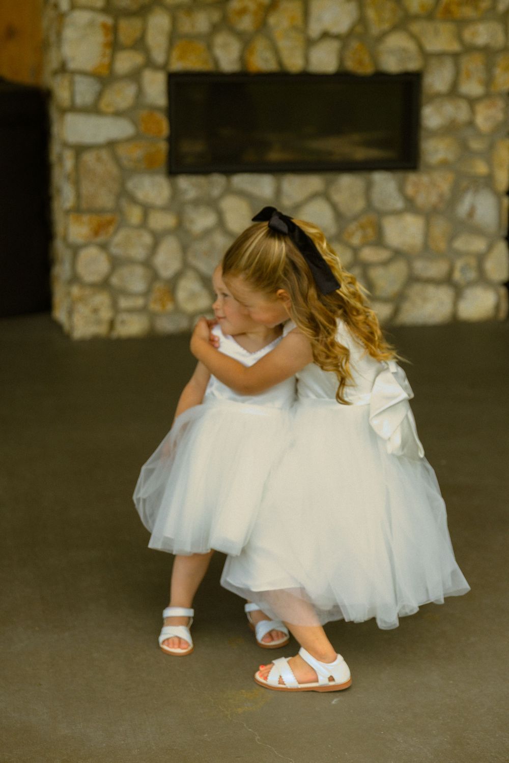 Two flower girls in white tulle dresses and sandals share a sweet hug near a stone fireplace.