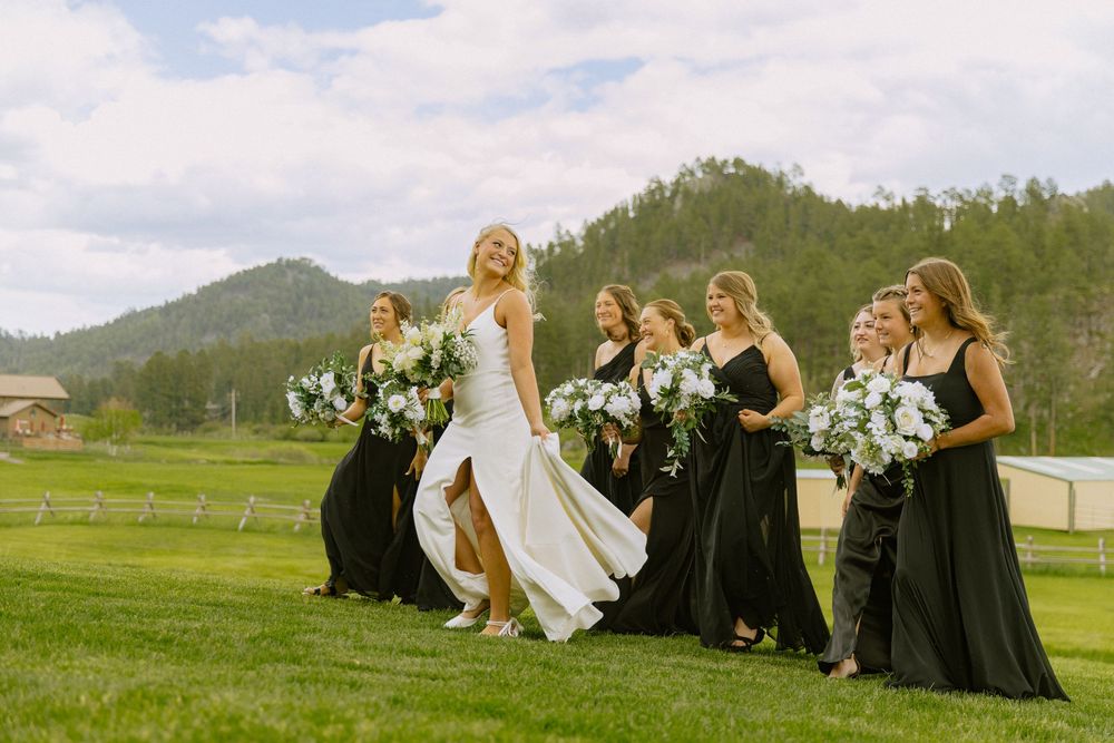 Bride in white gown walks with bridesmaids in black dresses holding white bouquets on green lawn with mountain backdrop.