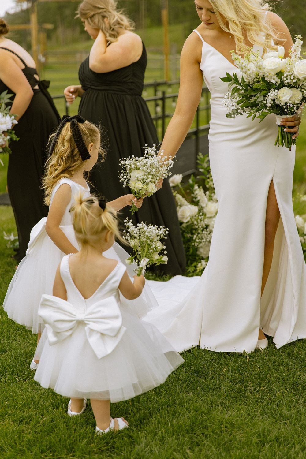 Bride in white gown holds bouquet with two flower girls in white dresses with bows on green lawn.